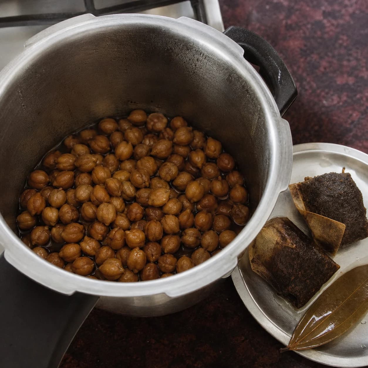 Tender deep-brown chickpeas in an open pressure cooker beside removed black tea bags and a bay leaf