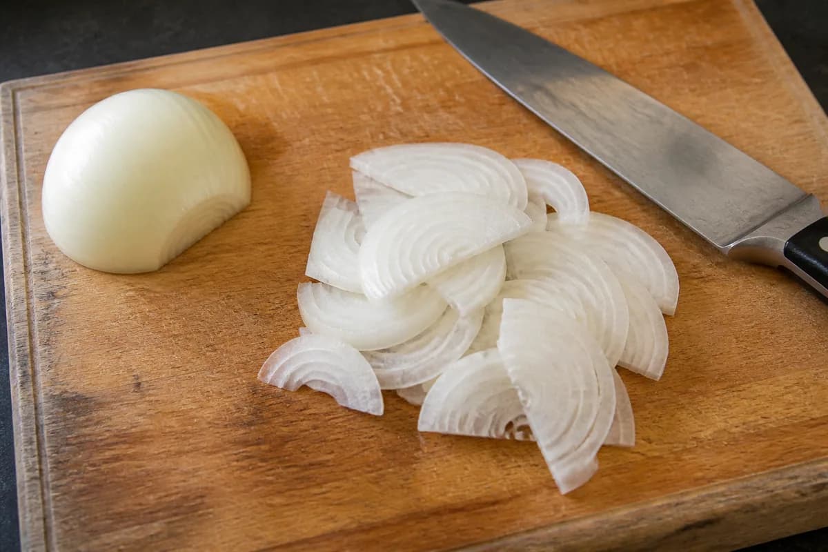A white onion sliced into thin half-moons on a wooden chopping board
