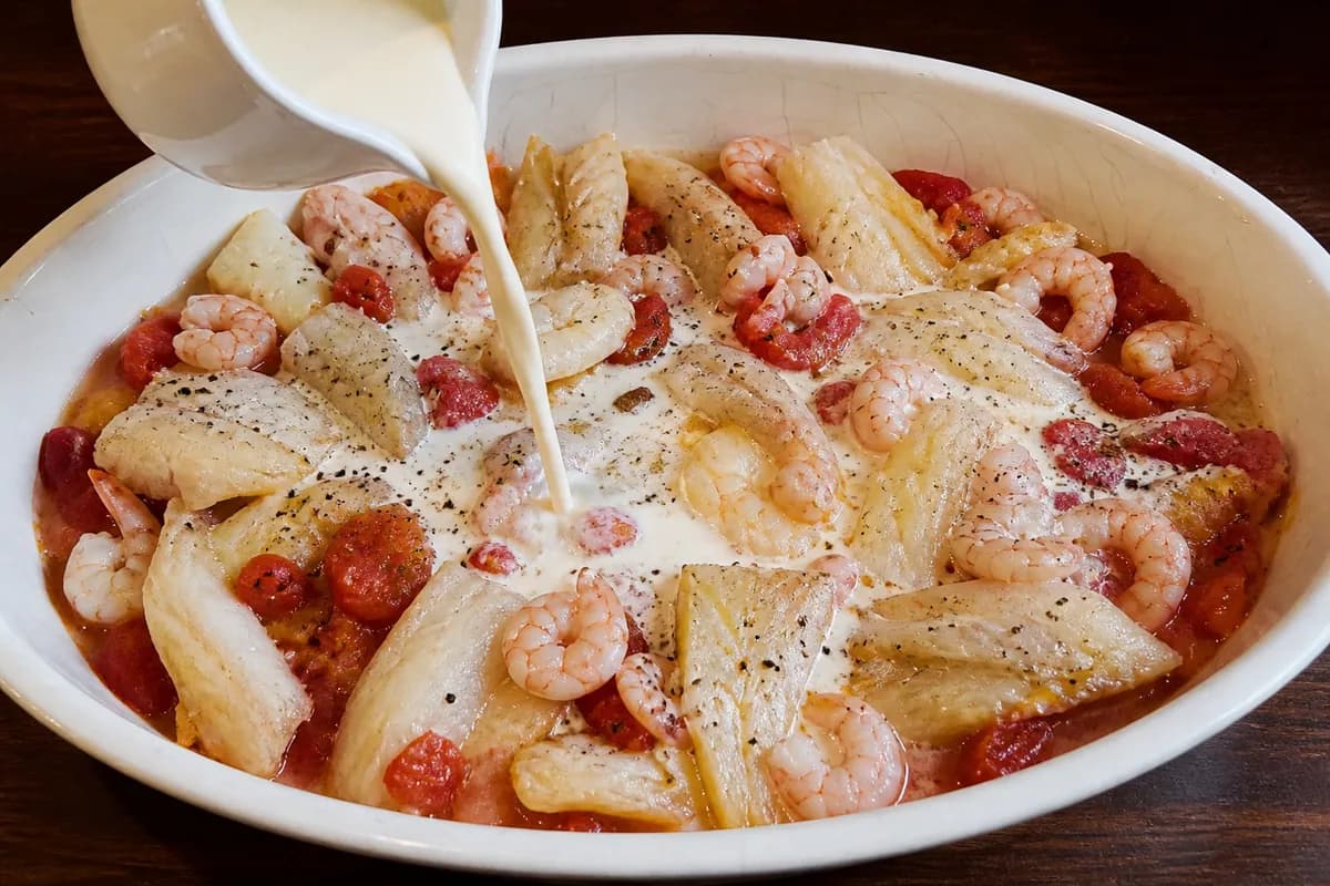 Milk and cream being poured over the haddock and tomato mixture in an oval baking dish